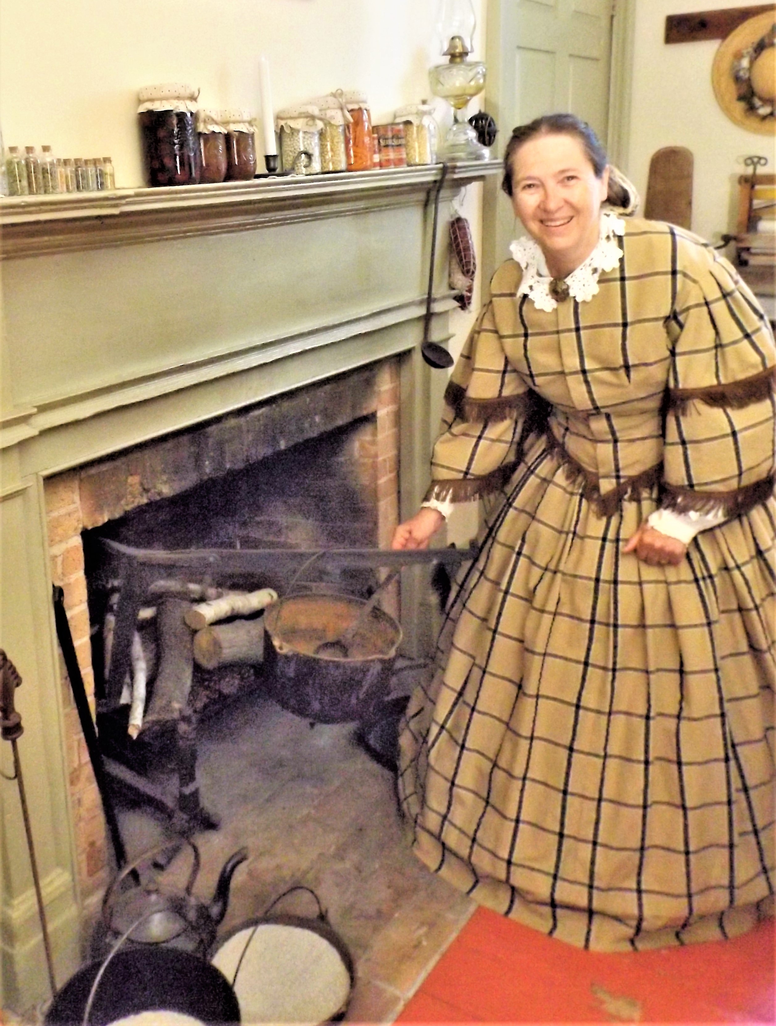 A woman in Victorian clothing demonstrates cooking over a hearth.