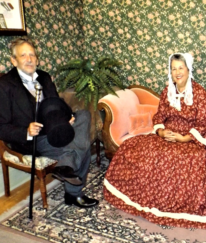 Two people in Victorian clothing sit in antique chairs, smiling at the camera.