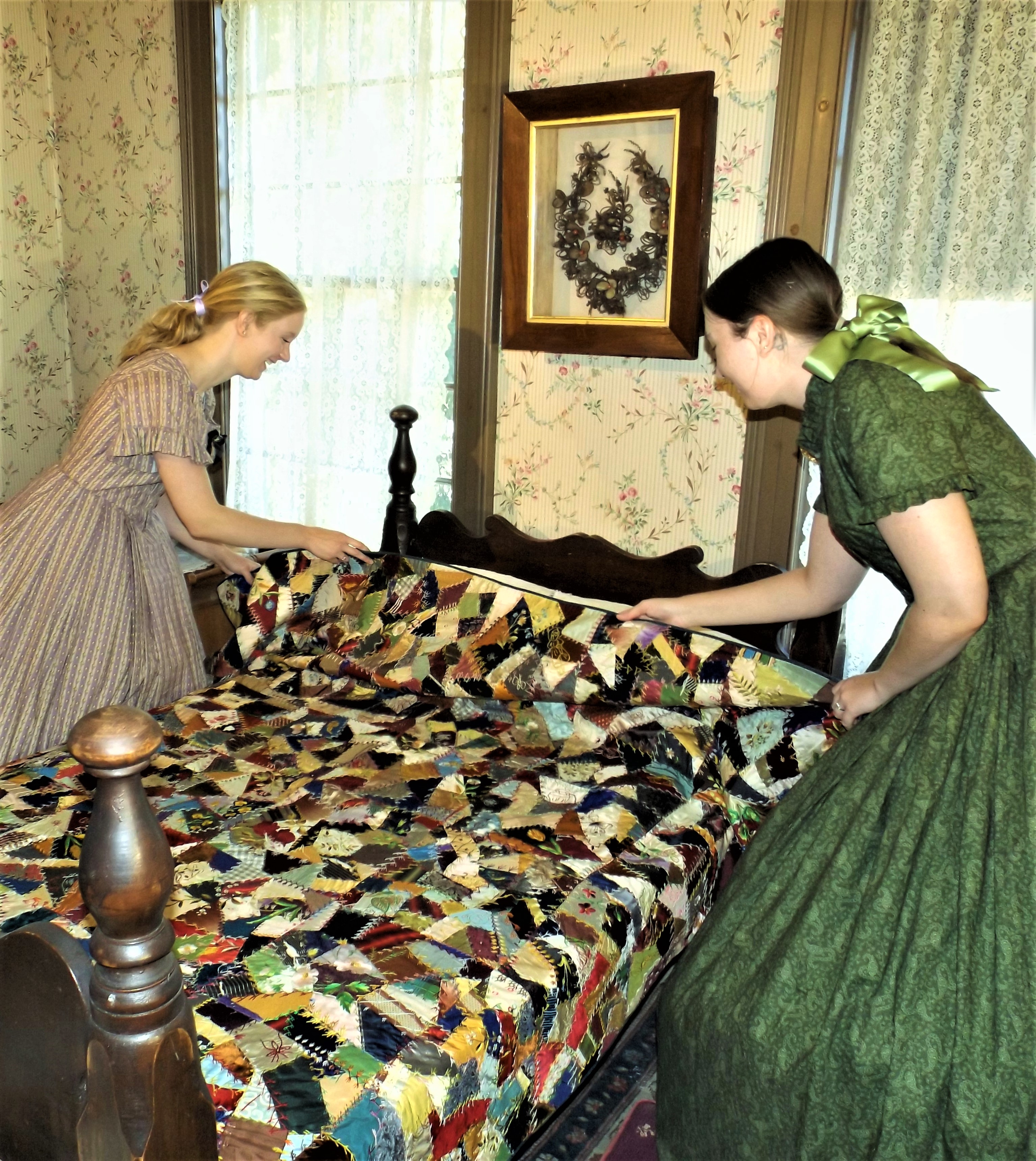 Two ladies in Victorian dresses lay an antique quilt on a bed.